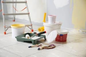 Painting supplies including brushes, rollers, buckets of paint, and a ladder leaning against a wall on a plastic sheet. There are several drips of paint on the plastic sheet floor.