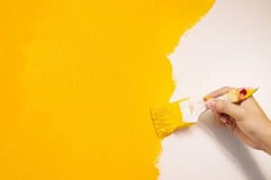 Close-up view of a hand carefully applying yellow paint to a wall with a paintbrush to avoid paint drips.