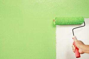 A person painting a house exterior wall with muted green exterior paint using a roller.