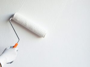 A gloved hand rolls white paint onto a freshly painted interior wall, showcasing the process of repainting interior walls.