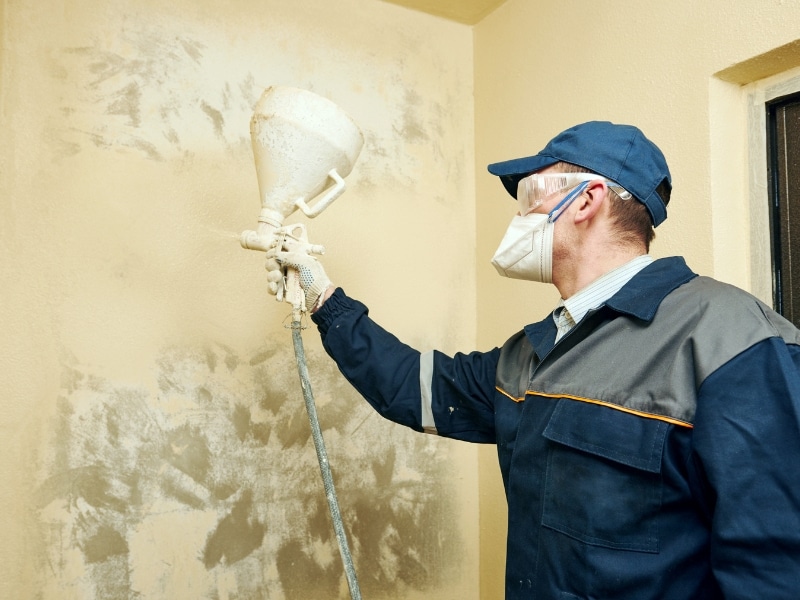 Professional worker using sprayer to spray paint walls indoors with protective gear.