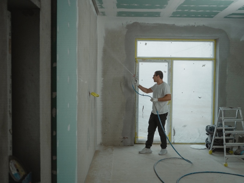 Worker using sprayer to spray paint walls indoors during home renovation.